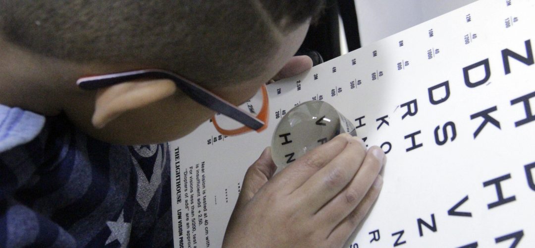 fotografia colorida de menino visto de cima. Ele usa óculos e está manuseando uma lupa de ampliação em uma tabela com letras diversas.