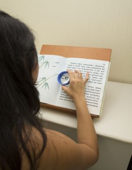 Fotografia colorida de uma mulher de cabelos compridos. Ela está de costas para a imagem, segura uma lupa de ampliação sobre um livro posicionado em uma mesa.