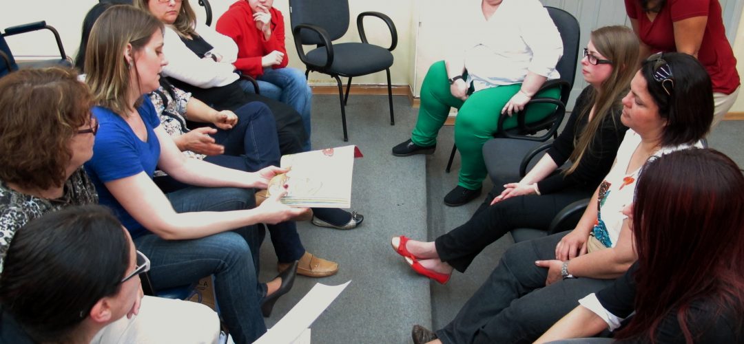 Fotografia colorida de cerca de 8 mulheres estão sentadas em círculo. Uma delas, à esquerda, segura um livro enquanto fala com as demais.