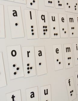 Descrição de imagem: fotografia de um grande painel, com fundo branco, em que há o alfabeto braille, em grande escalam, com as letras espalhadas por ele.