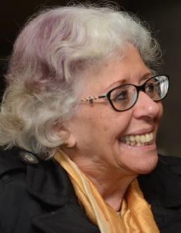 Descrição de imagem: Fotografia colorida de Marta Gil, levemente de perfil e sorrindo. Ela tem cabelos grisalhos e usa óculos.
